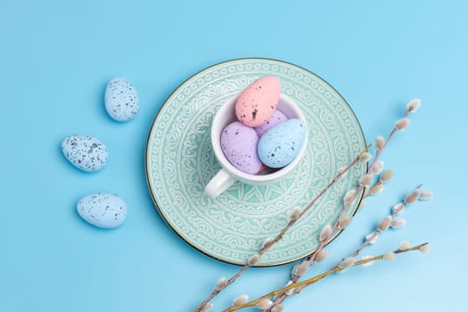 Porcelain cup with colored Easter eggs on a dish and willow branches with catkins on the blue background. Top view.