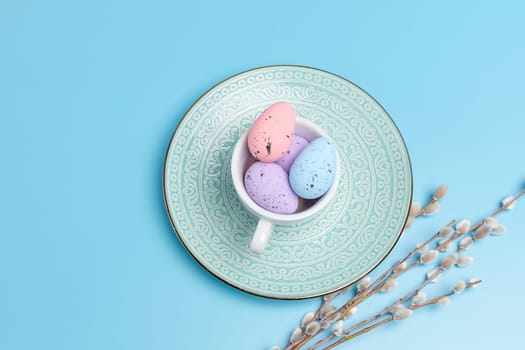 Porcelain cup with colored Easter eggs on a dish and willow branches with catkins on the blue background. Top view.