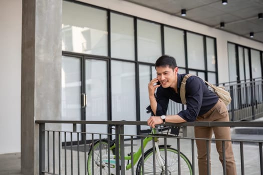 A young Asian businessman rides a bicycle to work. Standing outside the office talking on the phone and talking about business. Concept of reducing energy and reducing air pollution..