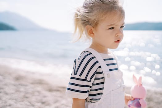 Little girl with a pink toy stands on the seashore and looks into the distance. High quality photo