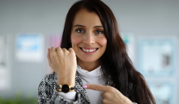 Close-up of smiling beautiful businesswoman pointing at wristwatch. Pretty young brunette woman in luxury costume. Time management and business company concept