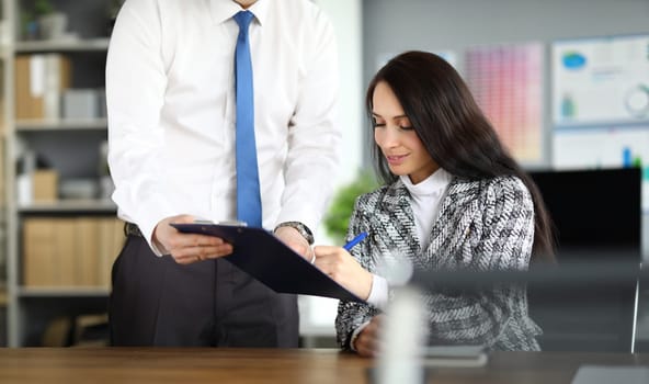 Portrait of businesswoman signing papers sitting in office. Attractive young woman wearing luxury suit. Male secretary holding clipboard. Business company and finance concept
