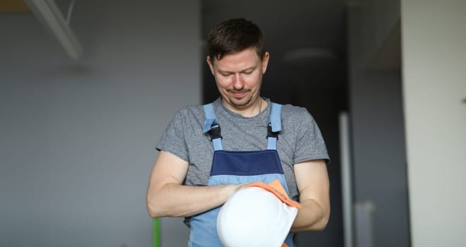 Portrait of handyman cleaning white protective helmet with rag. Room with grey walls. Disposable man in working uniform. Renovation and cleaner service concept