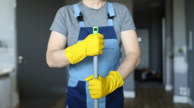 Close-up of professional cleaner holding mop. Male in special blue uniform and yellow protective gloves for work. Detergents for cleanliness. Cleaning service and renovation concept