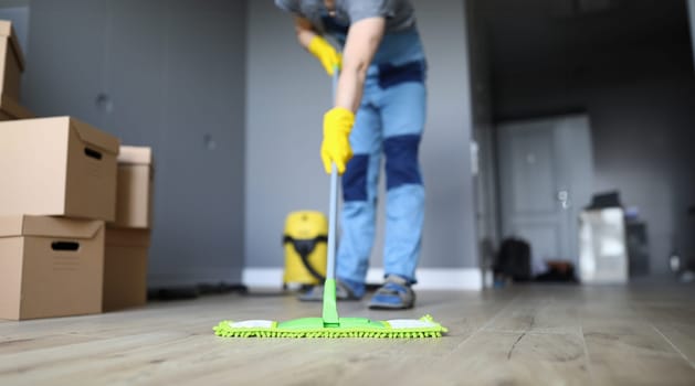 Close-up of professional cleaner washing floor with green mop. Man in working uniform. Handyman in gloves and cardboard boxes. Cleaning service and moving day concept