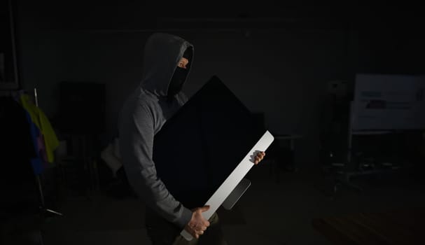 Portrait of man wearing grey hoodie and black mask. Male thief standing in dark room and holding expensive televisor from popular company. Stealth robbery concept