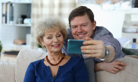 Portrait of cheerful mother and son taking selfie to remember. Smiling elderly woman posing for picture on sofa at home. Family relationship and spare time concept