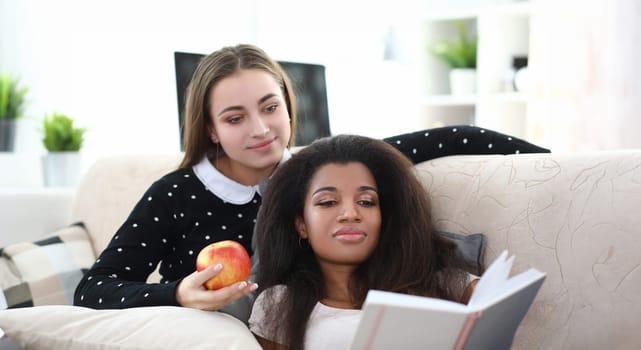Portrait of lovely girls friends spending time together reading book. Young woman sitting on sofa in living room holding apple fruit. Quality time and leisure concept