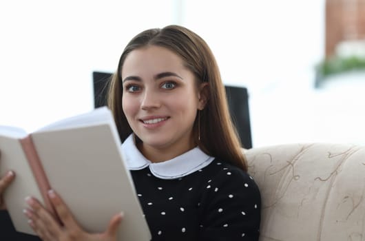 Portrait of beautiful young woman in cozy home interior sitting on sofa and reading personal diary. Charming smiling brunette holding notebook. Leisure concept