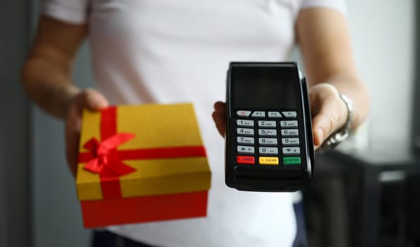 Close-up view of person holding delivery gift box with red bow and terminal for contactless payment for service. Quick credit card pay. Modern technology concept