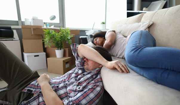 Portrait of exhausted husband and wife relaxing on cozy sofa in new purchased apartment. Stressed couple feel tired after moving. Unpacking boxes with stuff. Relocation concept