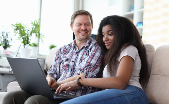 Portrait of couple sitting on sofa and choosing what film to watch. Afro-american young woman using laptop. Happy smiling middle-aged man. Relationship and quality time concept