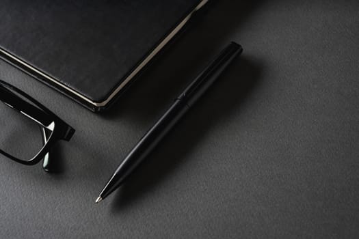 Top view of ballpoint pen, glasses and notepad lying on dark gray table