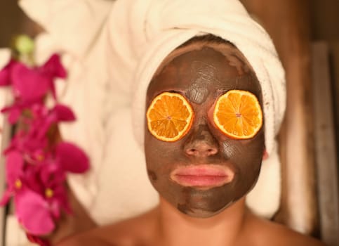 Top view of woman applying skin cleansing facial pack. Female person in mask with piece of orange on eyes on spa treatments. Beauty and relaxation concept