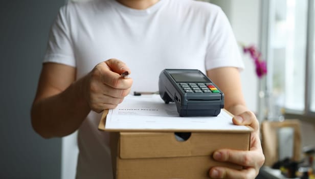 Close-up view of man holding cardboard box and payment terminal in hands. Credit card contactless paying for service. Package delivery and modern technology concept