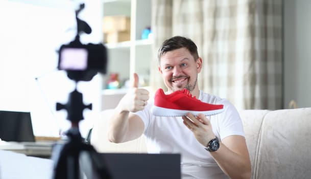 Portrait of smiling middle-aged man filming red sneaker on camera and tell in vlog. Male describing quality of new boots on display of digital device. Technology concept