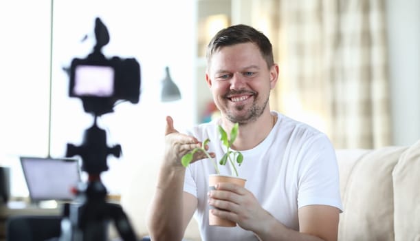 Portrait of middle-aged male smiling and showing on video camera plant in pot. Man beginning to film for vlogs. Beginner blogger and modern technology concept