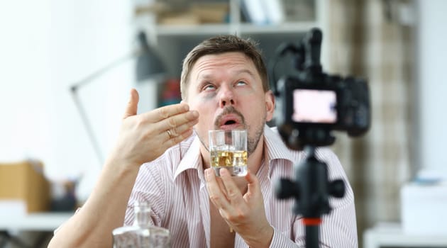 Portrait of drunk male filming on camera smelling glass of whiskey. Middle-aged man roll eyes and look messy. Tasting of alcoholic drink on videocamera. Blogger concept