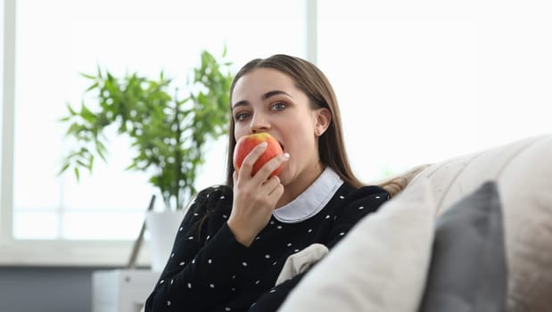 Portrait of attractive female biting juicy apple fruit. Cozy home interior. Pretty young woman eating vitamin sitting on sofa. Leisure and healthy food concept