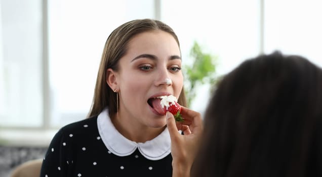 Close-up of afro-american lady feeding friend with juicy strawberry in whipped cream. Beautiful young brunette woman. Delicious dessert and spare time concept