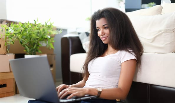 Portrait of good-looking afro-american woman using laptop and searching for information. on internet. Pretty young female sitting on floor in living room. Technology concept
