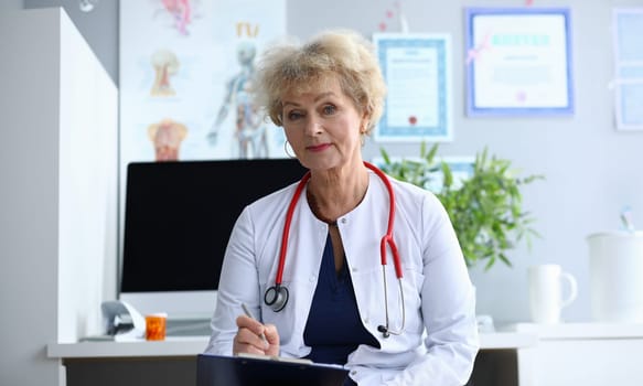 Portrait of smiling lady looking at camera with gladness. Cheerful elderly woman writing information about patient. Health care and prevention concept