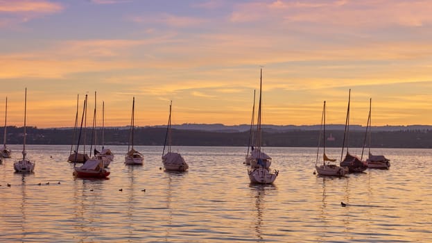 Bodensee Lake Sunrise Panorama. Morning Sunlight Over Tranquil Waters. Witness the mesmerizing dawn over Germany's Bodensee Lake, captured from a boat dock. Embrace the tranquil beauty of the early morning as the sun rises, casting a soft glow on the landscape. The peaceful scene features boats, yachts, and a charming water shack set against a backdrop of a captivating sky. Clouds delicately reflect on the calm water, creating a serene atmosphere. Immerse yourself in the serene beauty of a lakeside sunrise. Explore the harmony of nature, technology, and production as the day unfolds by the lake.