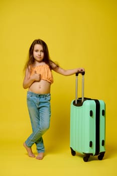 Full length vertical portrait of a Caucasian authentic baby girl, little child 6 years old in summer wear with suitcase, showing a thumb up, looking at camera, isolated over yellow studio background