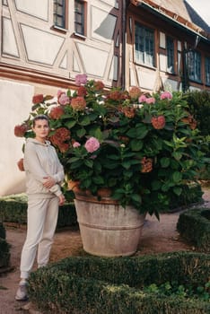 Attractive curly blonde woman walk on the city park street. Girl wear purple hoodie look happy and smiles. Woman make here me gesture standing near pink blooming bush flowers. Happy laughing girl