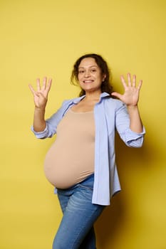 Smiling pregnant woman expecting a baby, showing nine fingers at the camera, isolated on yellow studio background. She's in last trimester of her happy pregnancy. Childbearing. Childbirth. Maternity.