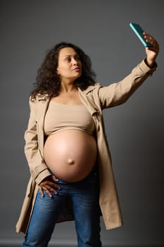 Stylish curly pregnant woman dressed in blue jeans and trendy trench, taking a selfie on mobile phone, sharing her lifestyle in last trimester of pregnancy on social media, isolated over gray backdrop
