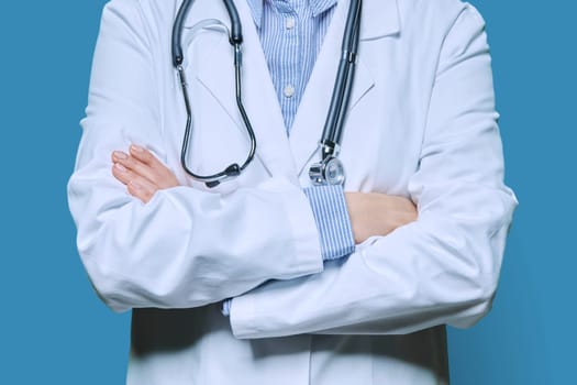 Medicine, medical services, health care concept. Close-up of crossed arms of confident female doctor in white lab coat with stethoscope on blue studio background