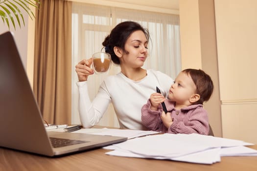 Cheerful businesswoman working at home with her little child girl and drinking coffee