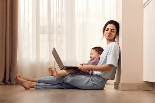 tired casual woman sitting on the floor and working on laptop with her little child girl at home