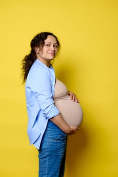 Side portrait of a happy pregnant woman putting hands on her big belly in last trimester of pregnancy, smiling looking at camera, isolated over yellow studio background. Maternity leave concept