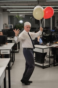 Portrait of a cheerful mature business man holding balloons in the office. Vertical photo