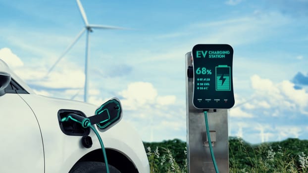 Electric car recharging energy from charging station by smart EV charger in wind turbine farm with nature outdoor background. Technological advance of alternative clean sustainable energy. Peruse