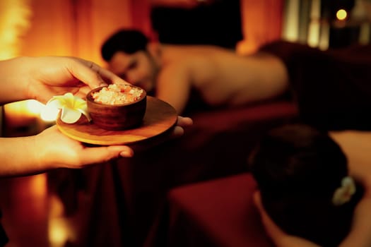 Masseur holding spa salt while couple customer having exfoliation treatment in luxury spa salon with warmth candle light ambient. Salt scrub beauty treatment in Health spa body scrub. Quiescent