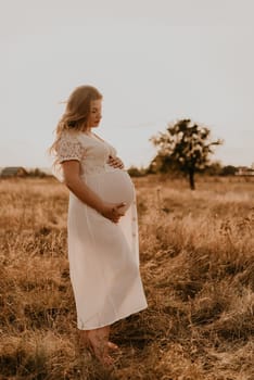 Caucasian pregnant young blonde woman in cotton white linen dress stand walking meadow on dry grass in summer at sunset nature. mother-to-be holds her hands on big round belly. tree in sunbeam