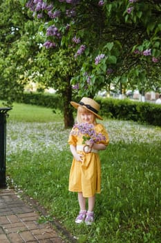 A little girl in a yellow dress and straw hat wearing a bouquet of lilacs. A walk in a spring park, blossoming lilacs