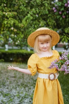 A little girl in a yellow dress and straw hat wearing a bouquet of lilacs. A walk in a spring park, blossoming lilacs