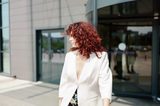 Portrait of a woman standing near a supermarket building. Caucasian model with long dark hair, wearing a white jacket and colored trousers