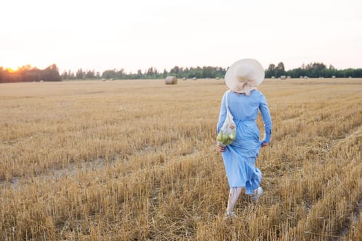 A red-haired woman in a hat and a blue dress walks in a field with haystacks