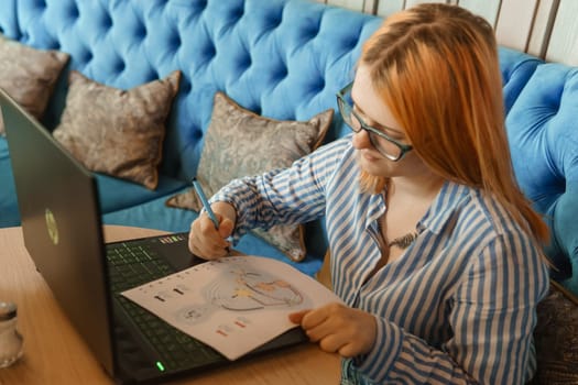 Tver, Russia-august 2, 2021. A woman in a cafe at a table with a laptop is studying the design of a person. The concept of studying esoteric sciences. A red-haired woman with glasses studies a rave card by human design.