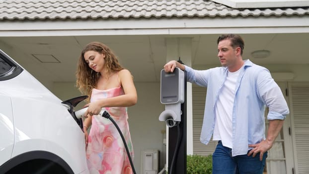 Happy and lovely couple with eco-friendly conscious recharging electric vehicle from EV charging station. EV car technology utilized as alternative transportation for future sustainability. Synchronos