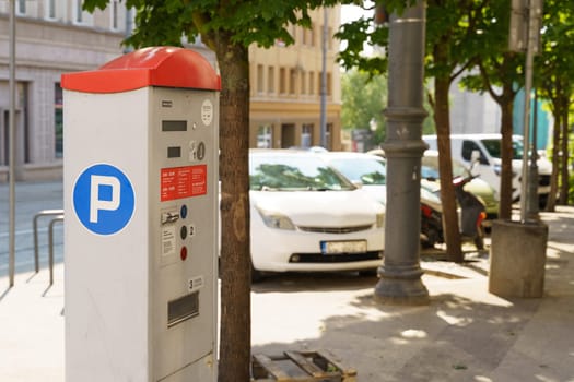 A parking meter stands on the sidewalk in a sunlit city, with cars park in the background and trees lining the street.