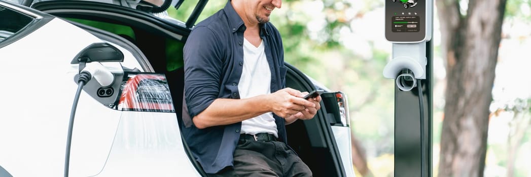 Man using smartphone online banking application to pay for electric car battery charging from EV charging station during vacation holiday road trip at national park or summer forest. Panorama Exalt