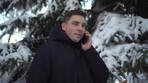 A young man speaks on the phone while standing against a background of spruce in winter. A man with a phone against a background of snowy spruce branches. 4k