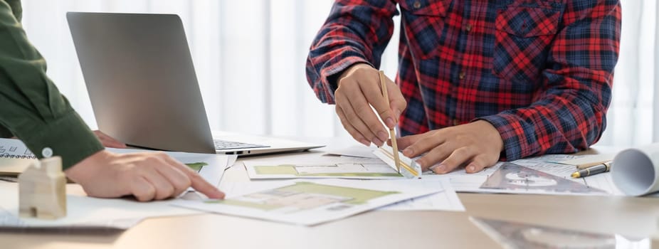 A portrait of skilled architect engineer hand writing a detail of blueprint during male engineer searching data from laptop at modern architectural office. Focus on hand. Closeup. Delineation.