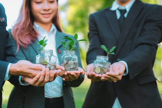 Concept of sustainable money growth investment with glass jar filled with money savings coins with businesspeople as eco-friendly financial investment nurtured with nature and healthy retirement. Gyre